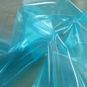 May include: A close-up of translucent, light blue plastic sheeting. The material is folded and layered, creating a dynamic interplay of light and shadow. The plastic has a glossy, reflective surface.