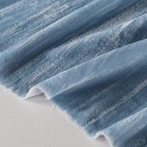 May include: Close-up of a soft, blue velvet fabric with a textured surface. The fabric is folded, revealing a white backing. The color gradient shifts from light to dark blue, creating a sense of depth and dimension. Ideal for apparel or home decor.