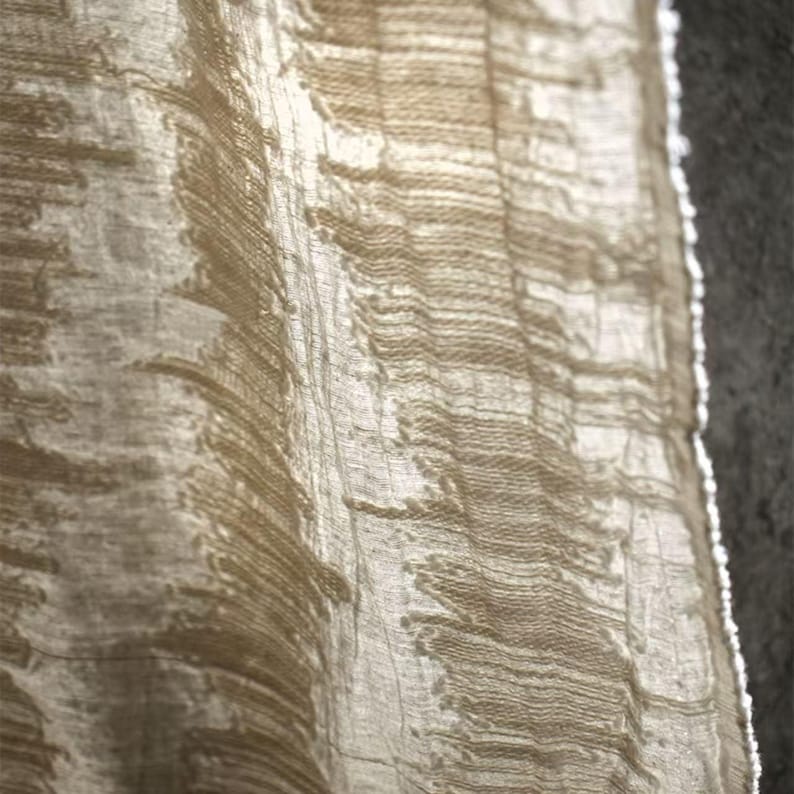 May include: Close-up of a textured, beige fabric with a layered, organic pattern. The fabric appears to be semi-sheer, with light filtering through, creating highlights and shadows. The material has a natural, raw aesthetic.