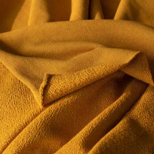 May include: Close-up of a folded mustard-yellow fabric. The fabric has a textured, woven appearance on one side and a softer, looped texture on the other. The folds create shadows and depth, highlighting the material's drape.