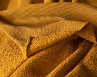 May include: Close-up of a folded mustard-yellow fabric. The fabric has a textured, woven appearance on one side and a softer, looped texture on the other. The folds create shadows and depth, highlighting the material's drape.