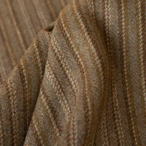 May include: Close-up of a textured fabric with a brown and tan striped pattern. The fabric has a woven appearance with vertical stripes of varying widths and colors. The material appears to be soft and slightly wrinkled, suggesting it is a natural fiber.