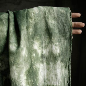 Puede incluir: Un trozo de tela verde con un dise&ntilde;o tie-dye. La tela presenta una mezcla de tonos verdes claros y oscuros, con detalles en blanco. Los bordes de la tela est&aacute;n deshilachados. La tela est&aacute; sujeta por una mano.