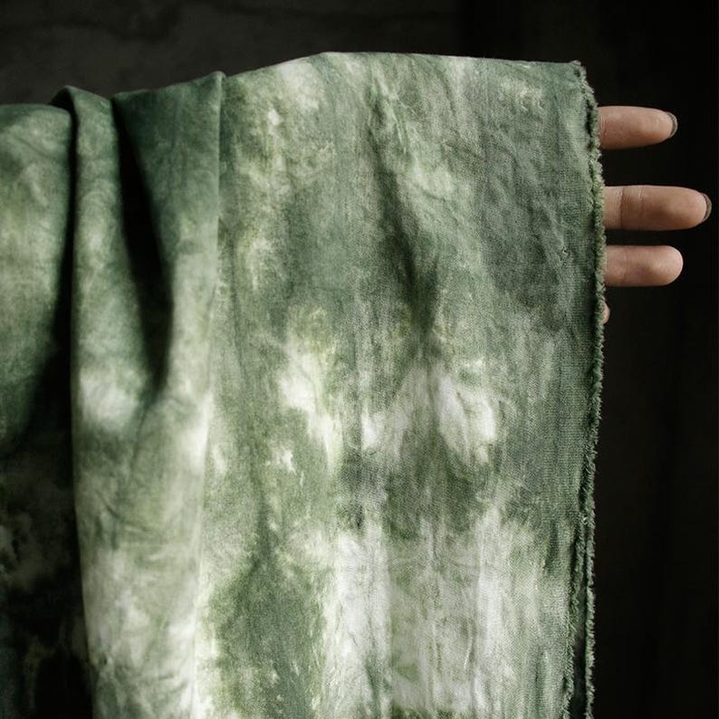 Puede incluir: Un trozo de tela verde con un dise&ntilde;o tie-dye. La tela presenta una mezcla de tonos verdes claros y oscuros, con detalles en blanco. Los bordes de la tela est&aacute;n deshilachados. La tela est&aacute; sujeta por una mano.