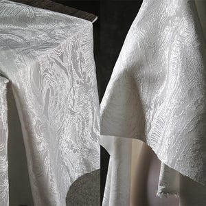 K&ouml;nnte beinhalten: Nahaufnahme eines wei&szlig;en Stoffes mit einem wirbelnden, strukturierten Muster. Der Stoff ist drapiert, mit Falten und Schatten, die Tiefe verleihen. Das Design &auml;hnelt einem marmorierten oder wasserartigen Effekt und erzeugt einen luxuri&ouml;sen Look. Der Stoff wird wahrscheinlich f&uuml;r Kleidung oder Heimdekoration verwendet.