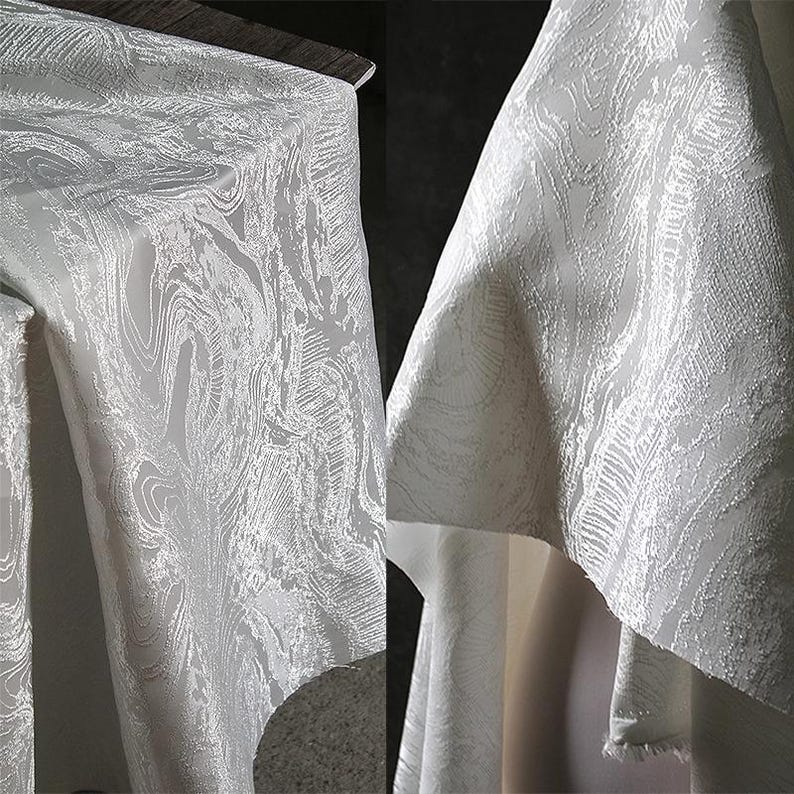 K&ouml;nnte beinhalten: Nahaufnahme eines wei&szlig;en Stoffes mit einem wirbelnden, strukturierten Muster. Der Stoff ist drapiert, mit Falten und Schatten, die Tiefe verleihen. Das Design &auml;hnelt einem marmorierten oder wasserartigen Effekt und erzeugt einen luxuri&ouml;sen Look. Der Stoff wird wahrscheinlich f&uuml;r Kleidung oder Heimdekoration verwendet.