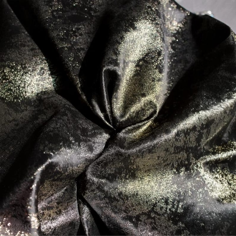 Puede incluir: Primer plano de una tela negra con un patr&oacute;n moteado dorado met&aacute;lico. La tela est&aacute; doblada, creando sombras y reflejos que enfatizan su textura y brillo. El material parece suave y fluido.