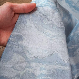 May include: A close-up of a fabric with a marbled design. The fabric is a blend of light blue, white, and gray, with a textured surface. The pattern resembles swirling clouds or water, creating a soft, flowing effect. The fabric is being held.