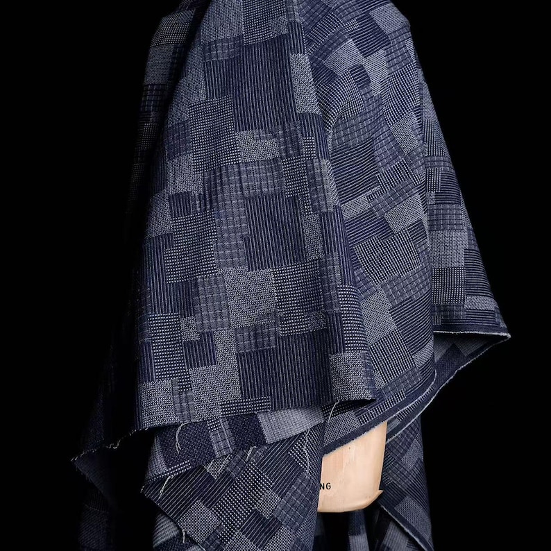 May include: A dark blue fabric featuring a patchwork design of various geometric patterns including stripes, grids, and small dots. The textile is draped over a light-colored mannequin form, showcasing the intricate woven textures and deep indigo tones.