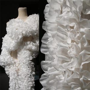 May include: A white garment with layered ruffles, displayed on a mannequin. The fabric appears sheer and delicate, with a voluminous, textured design. The ruffles create a cascading effect, suggesting a formal or artistic style.