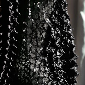 May include: Close-up of a textured black fabric with a complex, three-dimensional pattern. The fabric appears to be layered, creating a rough, uneven surface. The lighting highlights the intricate details and shadows of the material.