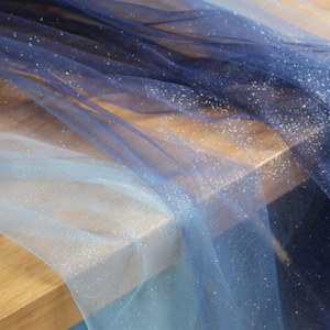 Pode incluir: Um tecido leve e transparente com um gradiente ombré que transita de um azul marinho profundo para um azul gelo pálido. O material é coberto com um acabamento de glitter prateado fino que capta a luz em toda a sua superfície translúcida.