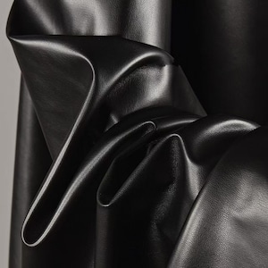 May include: Close-up of black leather fabric with a smooth, shiny surface. The material is draped and folded, creating shadows and highlights that emphasise its texture. The image showcases the supple quality of the leather.