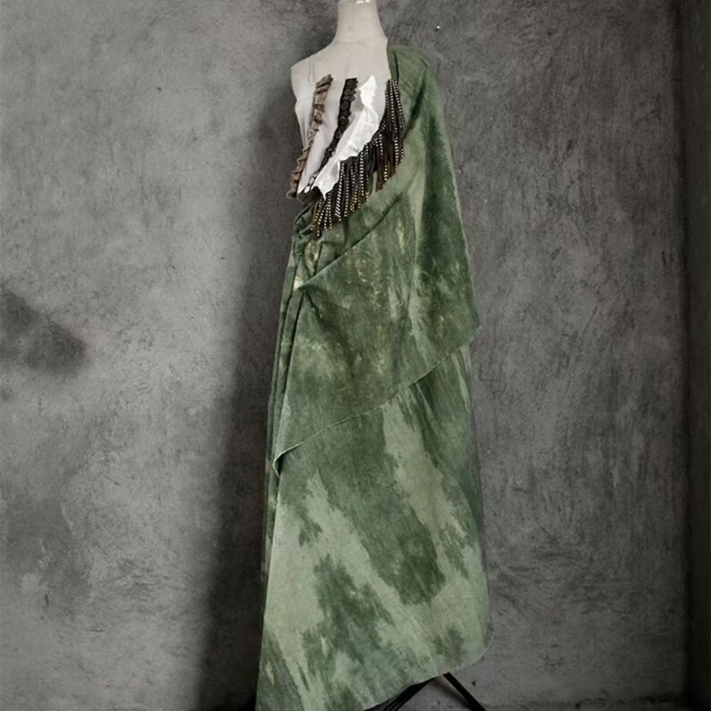 Puede incluir: Vestido verde drapeado sobre un maniqu&iacute;. El vestido tiene un dise&ntilde;o en capas con un estampado tie-dye. El corpi&ntilde;o presenta elementos decorativos blancos y marrones con flecos colgantes. Fondo gris.