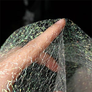 May include: Close-up of a shimmering, iridescent net fabric. The delicate mesh is held by a hand, showcasing the fine detail and light-catching qualities of the material. The fabric has a subtle, pearlescent sheen.