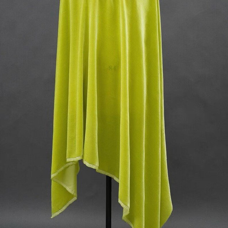 May include: A lime green velvet fabric draped over a black stand. The fabric has a soft, flowing texture with visible vertical folds. The edges are finished with a white stitch. The background is a dark gray.