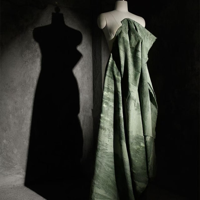 Puede incluir: Una tela verde oliva drapeada sobre un maniqu&iacute;. La tela tiene un aspecto veteado con acentos verdes claros y blancos. El fondo es oscuro, con la sombra de un maniqu&iacute; a la izquierda.