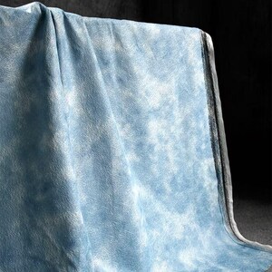 Op de afbeelding: Een lichtblauwe stof met een wit, wolkenachtig patroon. De stof is gedrapeerd, waardoor de textuur en het ontwerp zichtbaar worden. Het materiaal lijkt zacht en heeft een subtiele, gevlekte uitstraling.