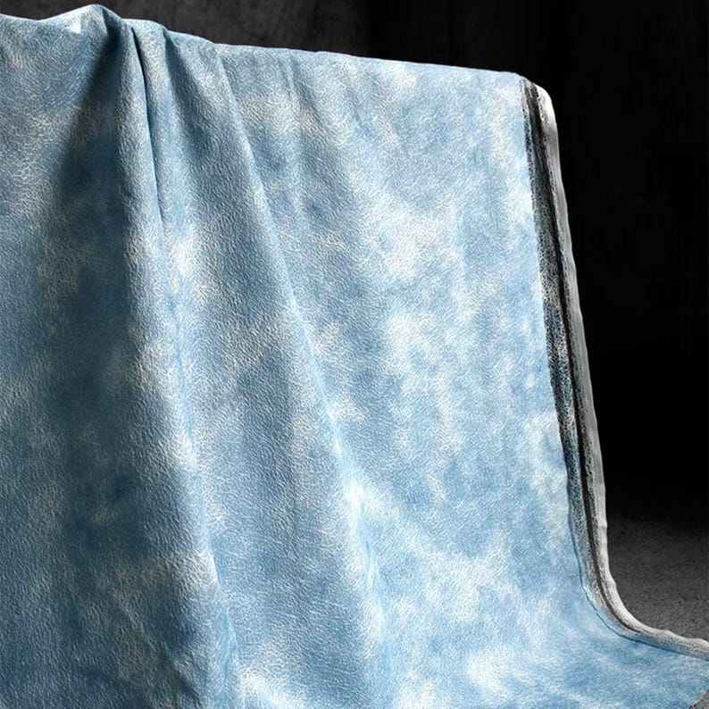 Op de afbeelding: Een lichtblauwe stof met een wit, wolkenachtig patroon. De stof is gedrapeerd, waardoor de textuur en het ontwerp zichtbaar worden. Het materiaal lijkt zacht en heeft een subtiele, gevlekte uitstraling.