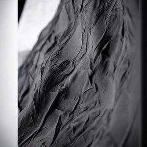 May include: Close-up of a dark gray fabric with a textured, rippled surface. The fabric appears to be draped, with folds and shadows creating a sense of depth and movement. The material has a matte finish.