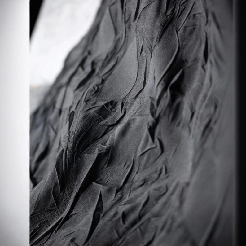 May include: Close-up of a dark gray fabric with a textured, rippled surface. The fabric appears to be draped, with folds and shadows creating a sense of depth and movement. The material has a matte finish.