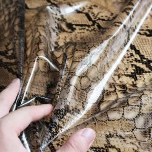 Könnte beinhalten: Ein flexibles Materialblatt mit einem realistischen Schlangenhautmuster in Beige-, Hellbraun- und Dunkelbrauntönen. Die Oberfläche hat ein glänzendes, reflektierendes Finish, das das Licht einfängt und die komplexe, überlappende Schuppenstruktur betont.