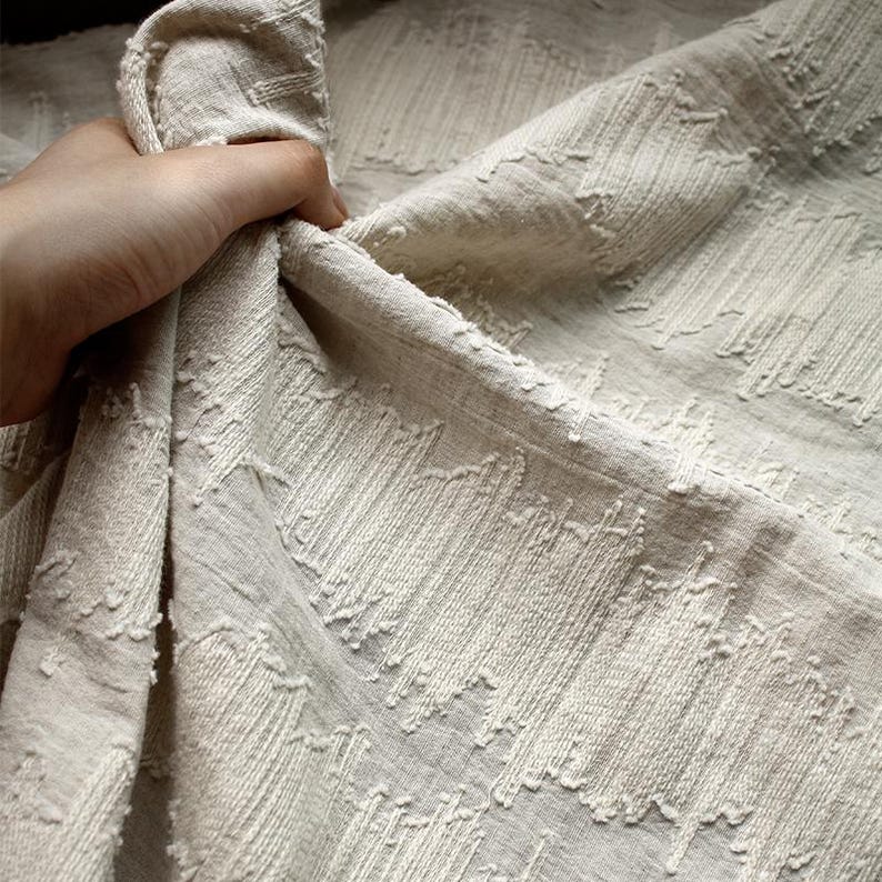 May include: Close-up of a beige fabric with a textured, woven pattern. The fabric has a slightly wrinkled appearance and is held in a hand, showing the drape and texture of the material. The weave creates a subtle, geometric design.