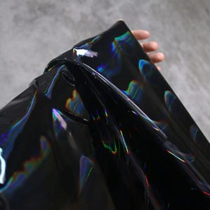 May include: A sheet of black, iridescent material. The material reflects a rainbow of colours, creating a shimmering effect. The material is held up, showing its flexibility and texture. The background is a neutral grey.