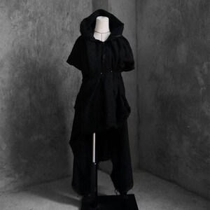 May include: A black hooded cape and matching pants set displayed on a mannequin. The cape features a button closure and a unique asymmetrical hemline. The pants have a distressed, ripped design. The outfit is made of a textured fabric.