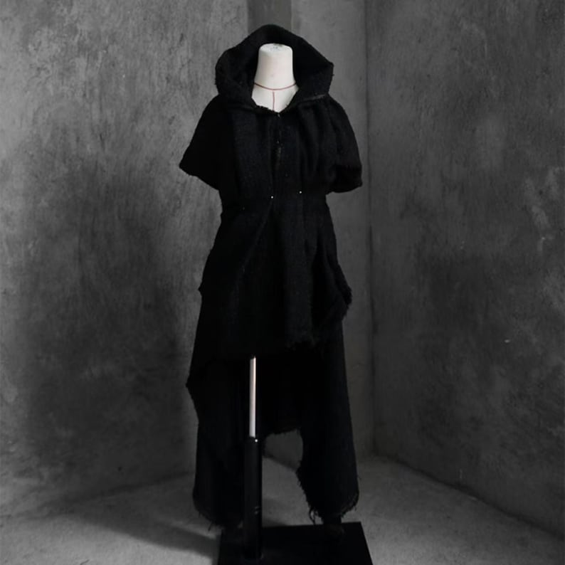 May include: A black hooded cape and matching pants set displayed on a mannequin. The cape features a button closure and a unique asymmetrical hemline. The pants have a distressed, ripped design. The outfit is made of a textured fabric.