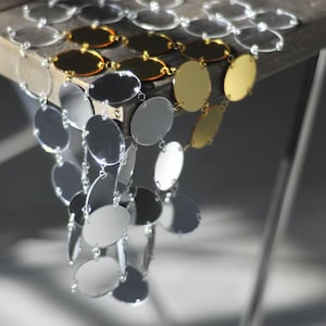 May include: A decorative hanging chain with silver, gold, and clear circular and oval reflective elements. The chain is suspended from a wooden surface, creating a cascading effect. The design incorporates various sizes and finishes of the reflective pieces.