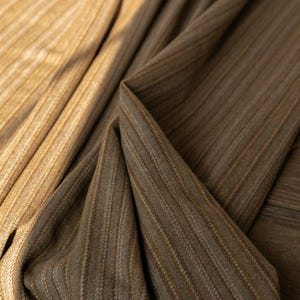 May include: Close-up of two fabrics with a vertical striped pattern. One fabric is a light tan color, and the other is a darker brown. The fabrics are draped, showing the texture and weave of the material. The image highlights the details of the textiles.