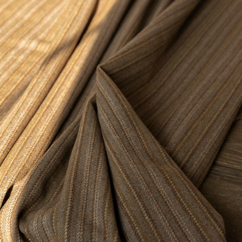 May include: Close-up of two fabrics with a vertical striped pattern. One fabric is a light tan color, and the other is a darker brown. The fabrics are draped, showing the texture and weave of the material. The image highlights the details of the textiles.