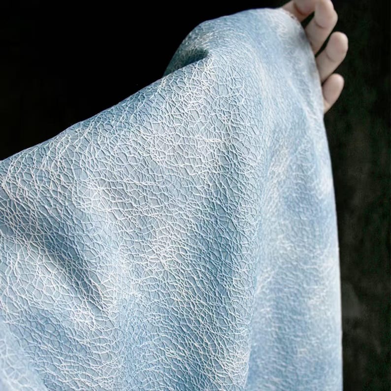 Op de afbeelding: Close-up van een lichtblauwe stof met een wit, webachtig patroon. De stof wordt omhoog gehouden, wat de textuur en de drape laat zien. De achtergrond is donker, waardoor de kleur en het ontwerp van de stof worden benadrukt.