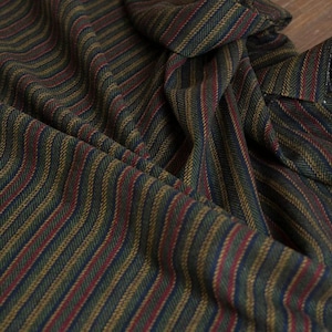 May include: Close-up of a folded fabric with a herringbone pattern. The fabric features vertical stripes in shades of green, gold, red, and blue. The material appears to be woven, with a textured surface and a slight sheen.