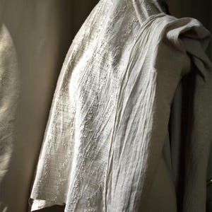 May include: A draped, off-white linen garment with a textured, woven appearance. The fabric has vertical and horizontal lines, creating a subtle pattern. The garment is draped over a form, showcasing its soft folds and natural texture.