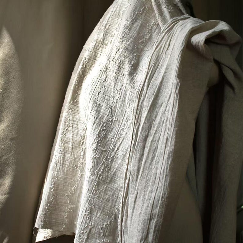 May include: A draped, off-white linen garment with a textured, woven appearance. The fabric has vertical and horizontal lines, creating a subtle pattern. The garment is draped over a form, showcasing its soft folds and natural texture.