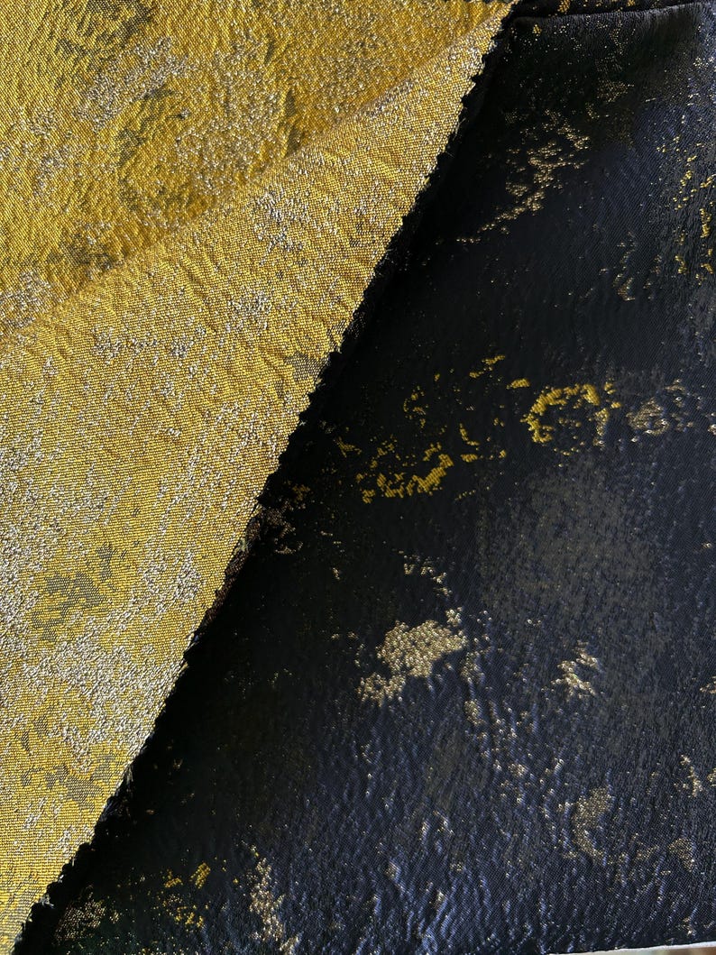 May include: Close-up of two fabric swatches. One is a textured gold fabric with a subtle pattern. The other is a black fabric with gold flecks, creating a contrasting visual. The fabrics are angled against each other.