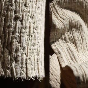 May include: Detailed view of off-white fabric with a textured surface. The left section has a quilted design with raised, uneven bumps. The right side shows a rippled texture with vertical lines and folds. The material looks natural, possibly linen or cotton.