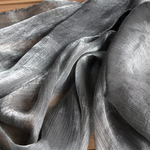 Peut inclure: Un tissu gris argenté translucide et chatoyant avec une texture froissée distincte. Le matériau présente un éclat métallique qui capte la lumière, créant des plis profonds et des vagues fluides sur la surface de ce textile léger et diaphane.