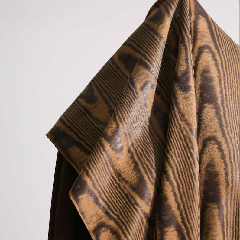 Puede incluir: Primer plano de una tela con un dise&ntilde;o de vetas de madera. La tela tiene un color base beige con l&iacute;neas curvas marr&oacute;n oscuro, creando una textura similar a la madera. La tela est&aacute; drapeada, mostrando sus pliegues y el contraste entre &aacute;reas claras y oscuras.
