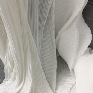 Op de afbeelding: Close-up van witte geplooide stof. De stof is gedrapeerd en gevouwen, waardoor een textuureffect met verticale lijnen ontstaat. Het materiaal lijkt zacht en licht, met subtiele variaties in toon en schaduw.