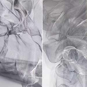 May include: Two-panel image featuring flowing, sheer fabric in shades of gray. The fabric is arranged in soft, undulating folds, creating a sense of movement and texture. The material appears to be a fine mesh or netting, with a subtle sheen.