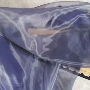 Può includere: Primo piano di un tessuto trasparente e fluido in tonalità blu e bianco. Il tessuto sembra essere organza o un materiale simile, con una lucentezza sottile e una trama delicata e ariosa. Le pieghe e gli strati creano una sensazione di movimento.
