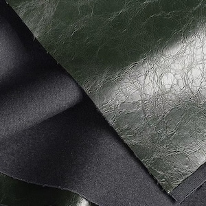 May include: Close-up of dark green leather fabric with a textured, wrinkled surface. The leather is folded over a layer of black fabric, revealing its underside. The image highlights the contrast between the two materials.