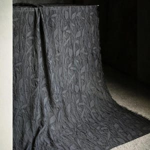 May include: Dark gray fabric with a leaf pattern draped over a table. The fabric has a textured appearance with raised leaf designs. The material cascades down, creating soft folds and shadows. The overall effect is elegant and textured.