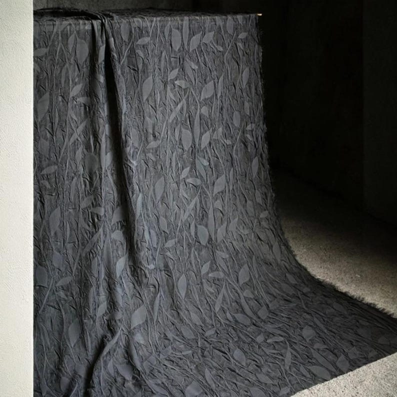 May include: Dark gray fabric with a leaf pattern draped over a table. The fabric has a textured appearance with raised leaf designs. The material cascades down, creating soft folds and shadows. The overall effect is elegant and textured.