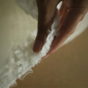 Op de afbeelding: Close-up van een witte, zachte stof met een delicate, gerafelde rand. De stof wordt door een hand vastgehouden, wat de zachte textuur en het potentieel voor textiel of ambachten benadrukt. De achtergrond is een warme, neutrale tint.