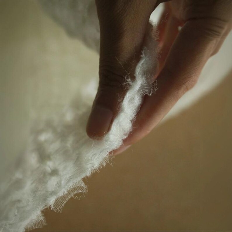 Op de afbeelding: Close-up van een witte, zachte stof met een delicate, gerafelde rand. De stof wordt door een hand vastgehouden, wat de zachte textuur en het potentieel voor textiel of ambachten benadrukt. De achtergrond is een warme, neutrale tint.