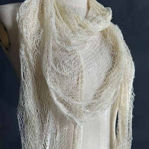 Könnte beinhalten: Cremefarbener Schal mit offenem Gewebe, drapiert auf einer Schaufensterpuppe. Der Schal hat eine zarte, netzartige Textur mit einem weichen, fließenden Aussehen. Der Stoff hat einen hellen, neutralen Ton, passend für verschiedene Outfits. Ein Modeaccessoire.